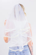 Person wearing a white veil with floral trim on a plain background